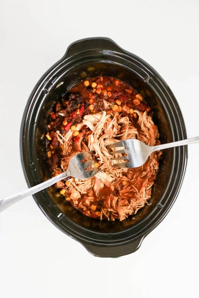 two forks in a crockpot with shredded chicken in a tomato sauce with beans and corn